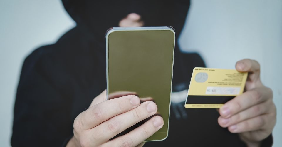 Photo d'une personne cagoulée tenant un smartphone et une carte de crédit, symbolisant la fraude en ligne et la cybersécurité.