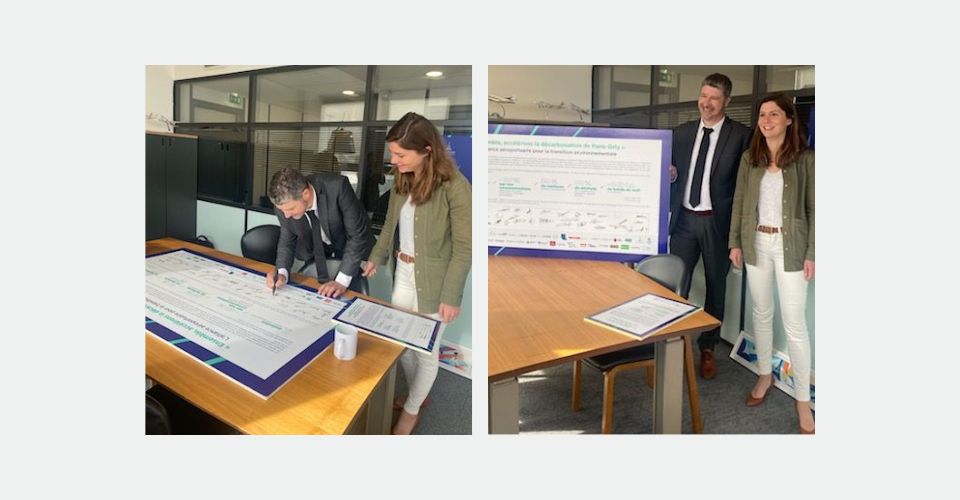 Deux personnes, un homme et une femme, debout à côté d'une table, regardant et signant un grand document.