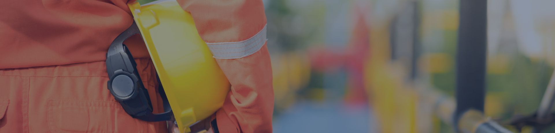 Un ouvrier de dos, en combinaison de travail orange, tenant un casque de chantier jaune sous le bras, marchant sur une plateforme industrielle.