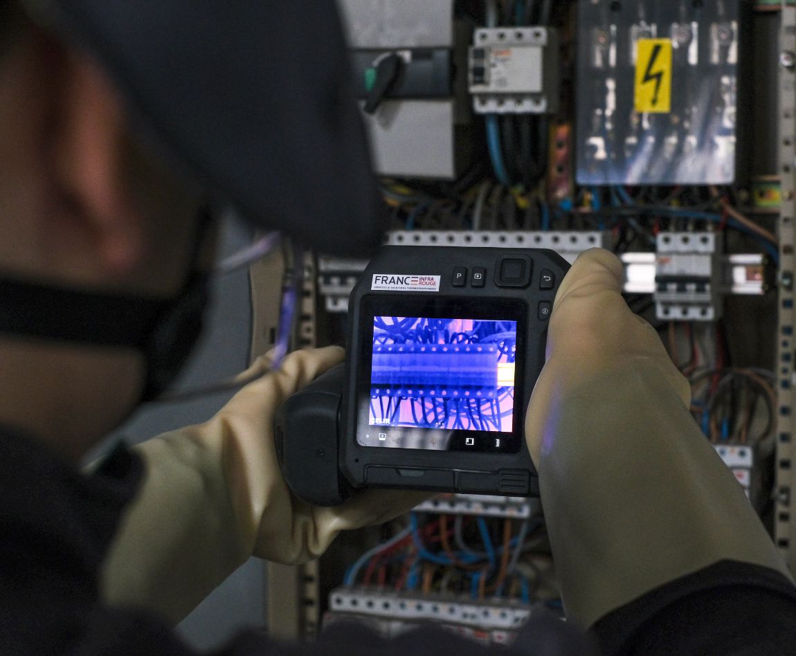Un technicien de dos, ganté, utilise une caméra thermique pour inspecter un tableau électrique. L'écran de la caméra montre une image en couleurs vives des composants internes.
