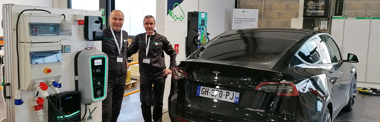 Deux collaborateurs Apave devant une voiture Tesla et un table