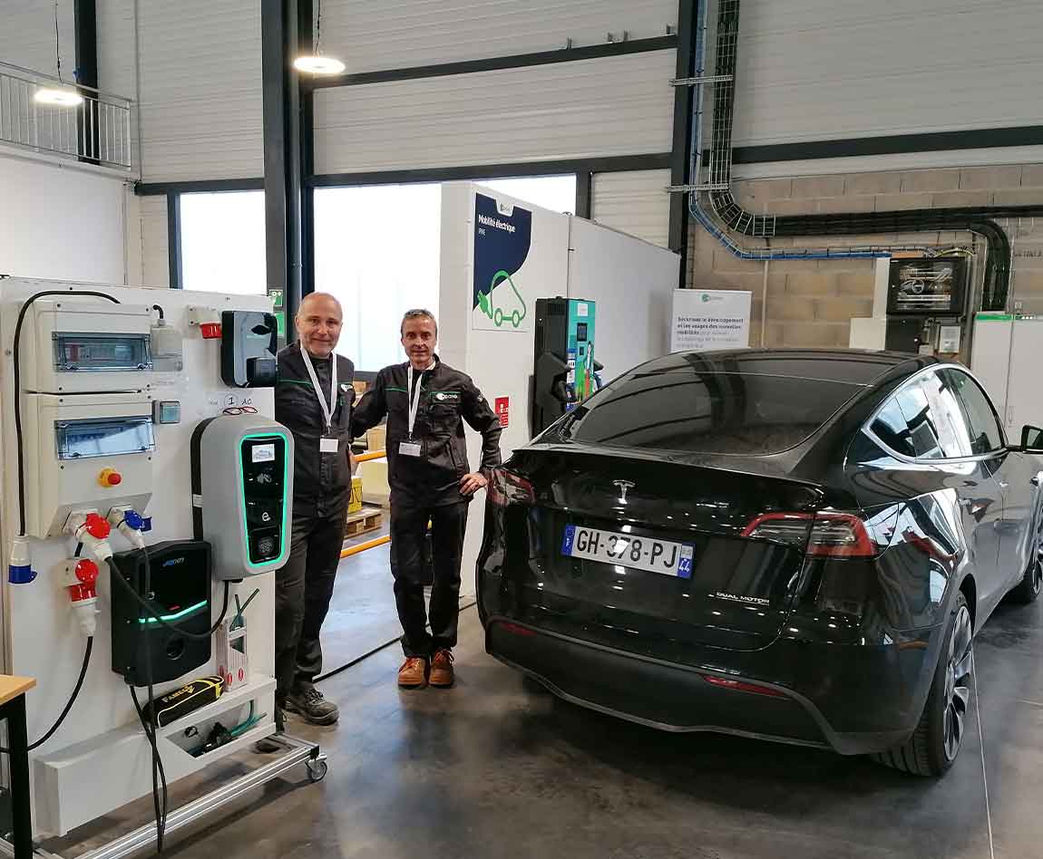 Deux collaborateurs Apave devant une voiture Tesla et un table
