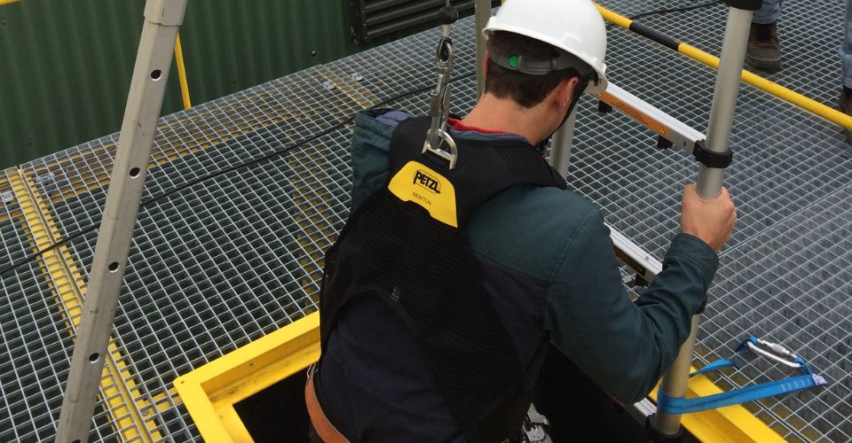 Un ouvrier avec un casque de chantier blanc et un harnais de sécurité est en train de descendre dans une trappe au sol.