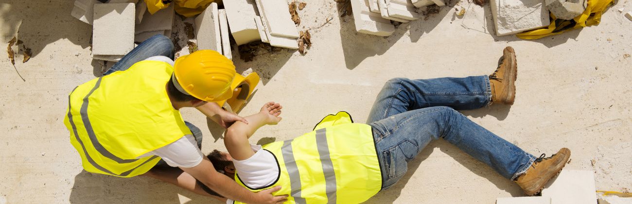 Vue en plongée d'un ouvrier blessé en gilet de sécurité jaune allongé au sol, secouru par un autre ouvrier.
