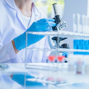 Une personne en blouse de laboratoire et gants bleus utilise un microscope avec des tubes à essai en arrière-plan.