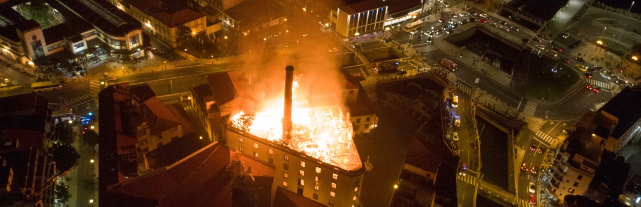 Vue aérienne nocturne d'un bâtiment industriel en flammes au cœur d'une zone urbaine, illustrant l'ampleur et la dangerosité d'un sinistre incendie majeur.