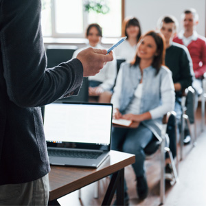 Un groupe de personnes suit une présentation en salle de classe, un homme au premier plan tenant un stylo devant un ordinateur portable.