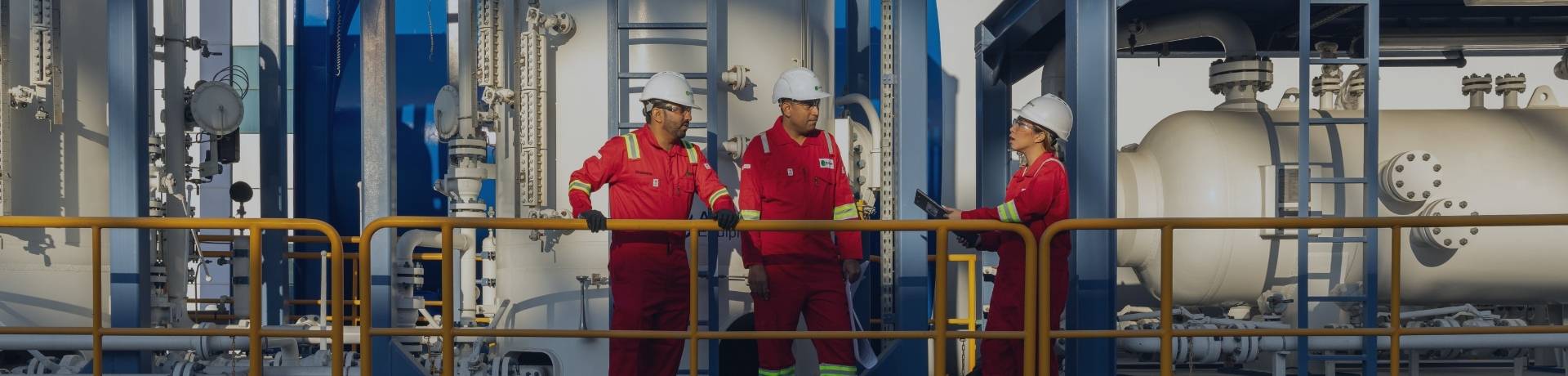 Three people in red jumpsuits and hard hats stand on a platform, inspecting large industrial equipment.