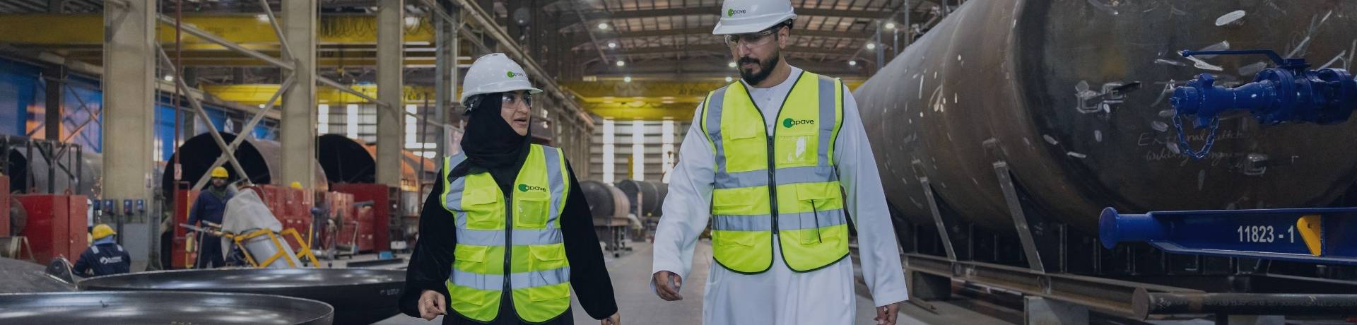  Two professionals wearing safety vests and hard hats walk through a manufacturing facility, discussing a management system.