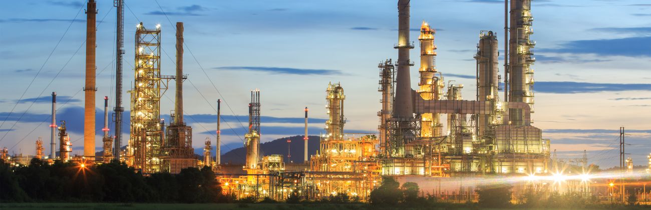 A night view of an illuminated oil refinery or petrochemical complex, with its chimneys and towers reflecting in the water.