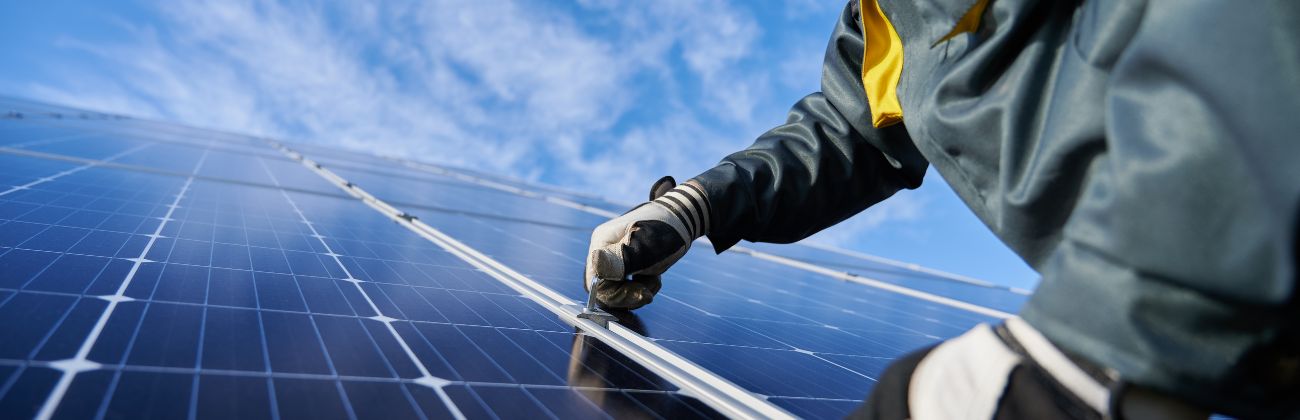 A technician wearing work gloves is inspecting and cleaning a row of solar panels against a blue sky with clouds.