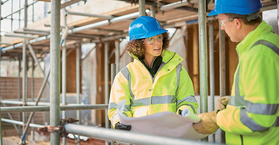 Un homme et une femme discutent sur un chantier de construction. Ils portent des manteaux jaunes réfléchissants et des casques bleus.
